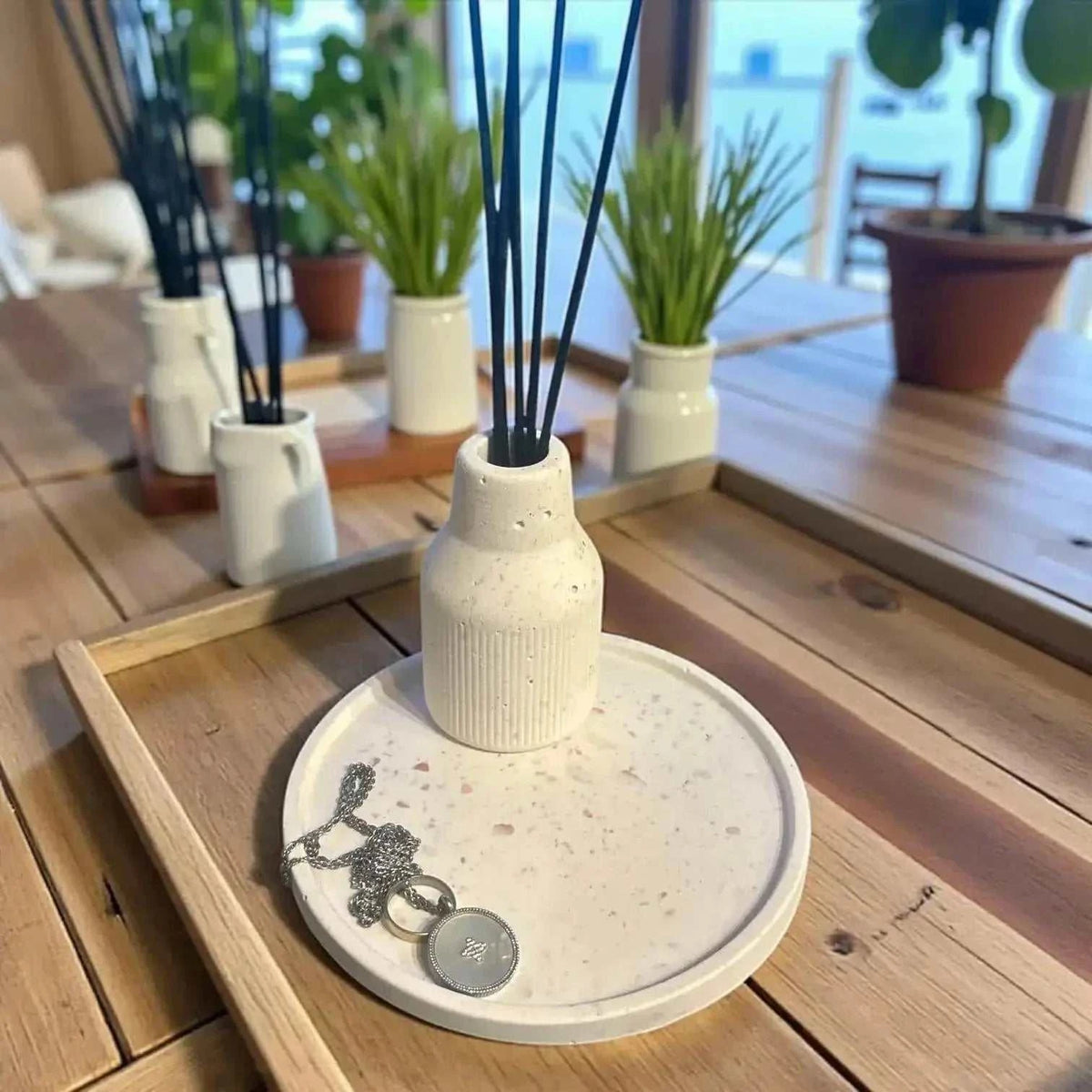 Diffuseur à Parfum en Jesmonite blanc crème avec rainures, posé sur un plateau rond terrazzo beige et marron contenant un pendentif argenté.